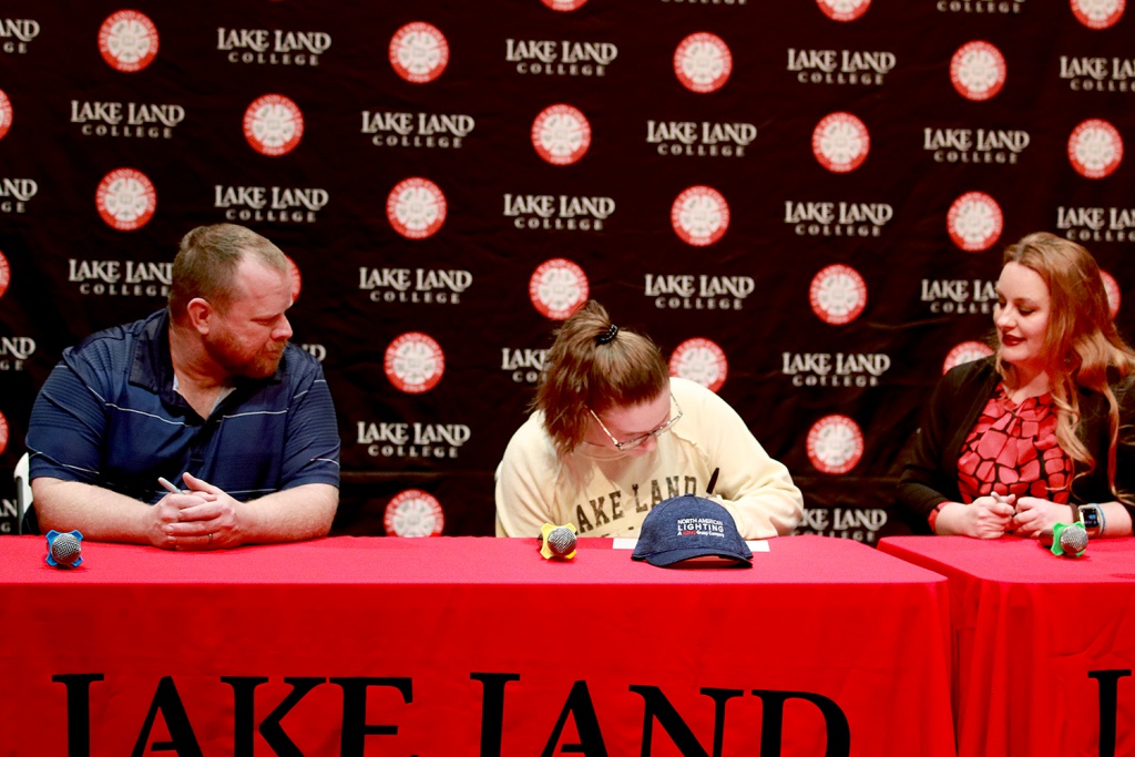 One man and two women sit at a table while the young woman in the middle is signing a paper.