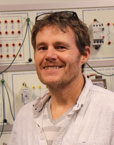 A photograph of Michael Beavers standing in an electronic lab and smiling at the camera.