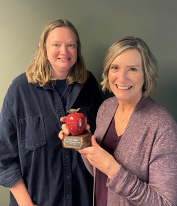 Two white women pose smiling. The woman on the right holds an engraved apple-shaped figurine.