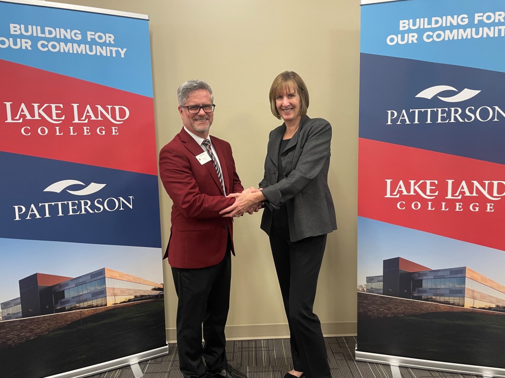 Lake Land College President Josh Bullock shakes hands with Vice President and General Manager of Dental Software Business at Patterson Companies Cecile Schauer. The two stand between large banners bearing the Lake Land College and Patterson logos as well as images of the building.