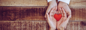 A photograph two larger hands encompassing two smaller hands on either side of a wooden table. The smaller hands are holding a red felt heart to the camera.