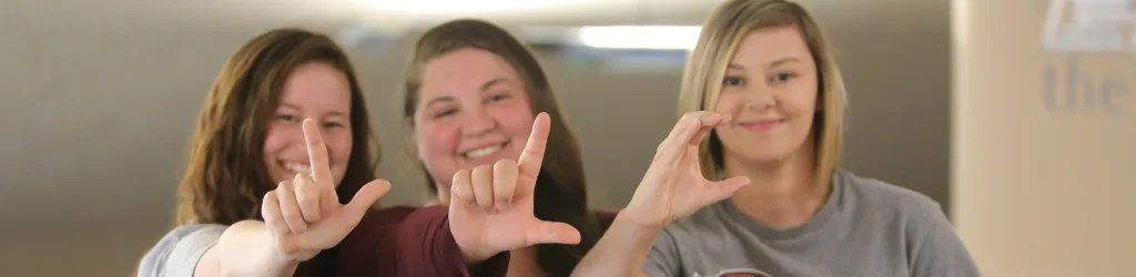 3 students standing together and signing L L C with their hand