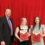 Students holding an award and posing with an instructor for a photo in front of a red curtain