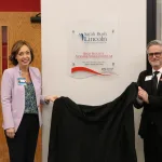 Two people holding a curtain and posing for a photo next to a sponsor sign for a new nursing simulation lab