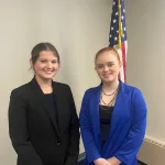 Two women posing for a photo inside in front of the American flag